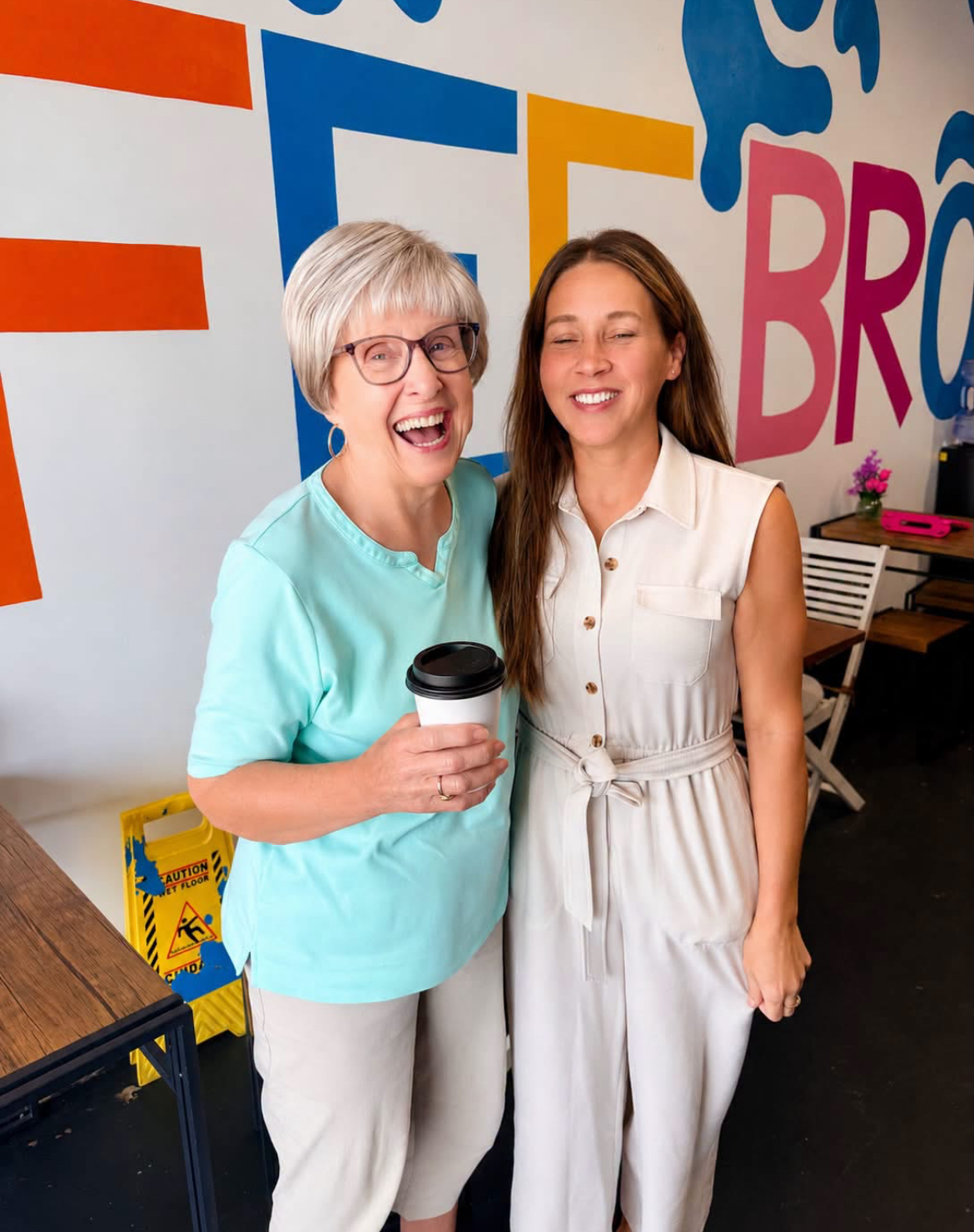 Two guests laughing with coffee in front of the Kofee Broz mural